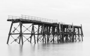 This black and white art print depicts an old, fading pier shrouded in morning fog, creating a timeless and mysterious atmosphere.